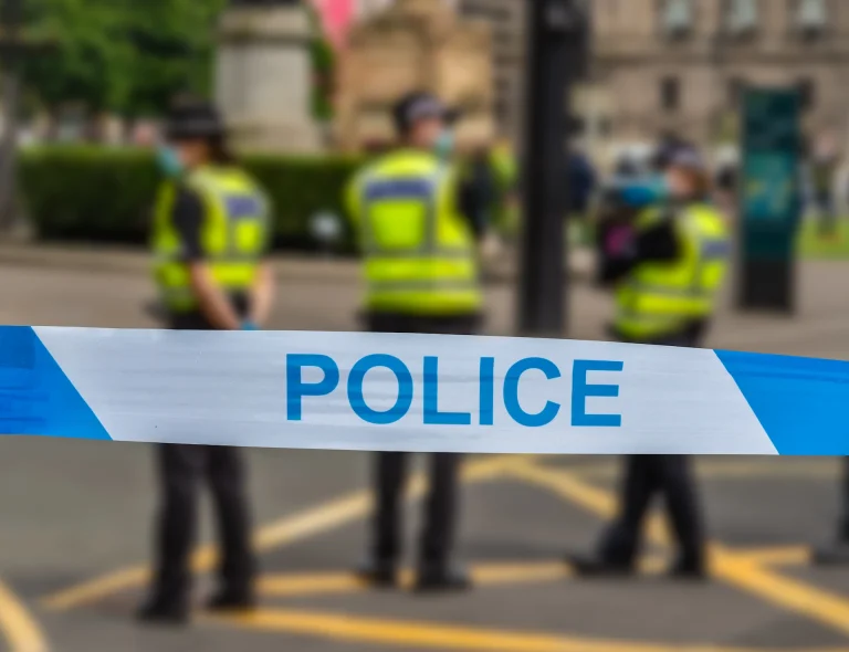 Police tape reading 'Police' blocks a street while officers in high-visibility vests stand behind the cordon.