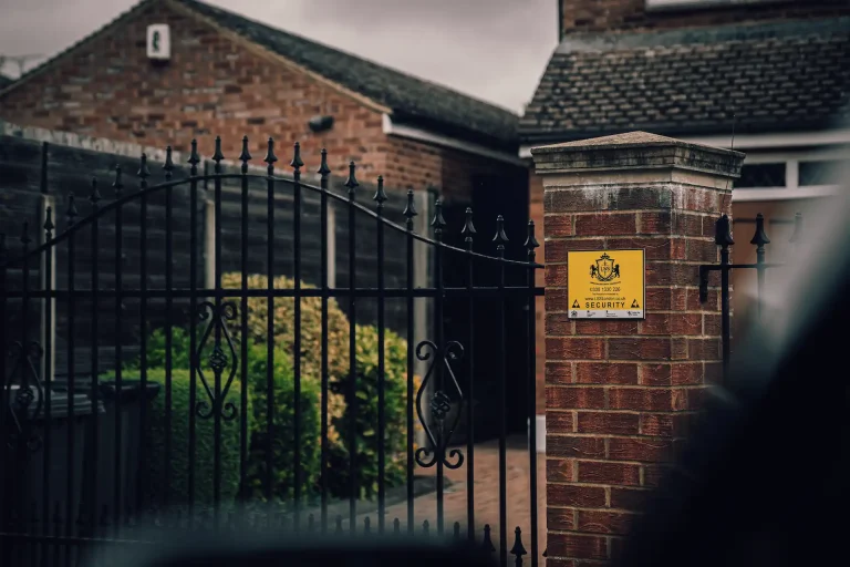 Black wrought-iron driveway gate and brick pillar with yellow LSS security sign, indicating monitored residential property.