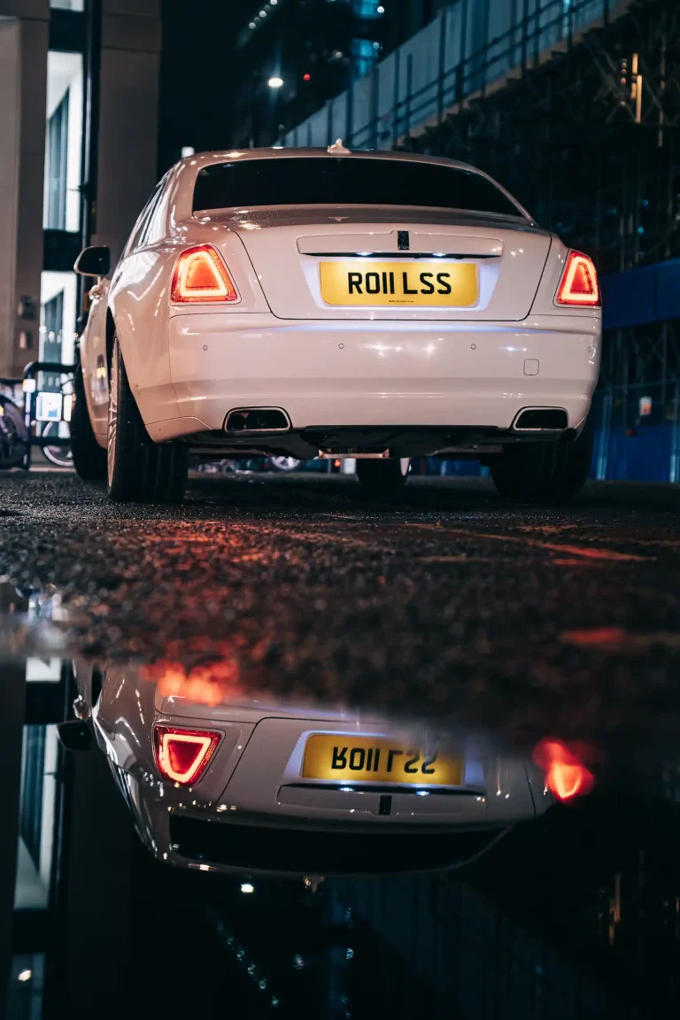 White Rolls-Royce parked on a wet city street at night, with rear lights reflected in a puddle.